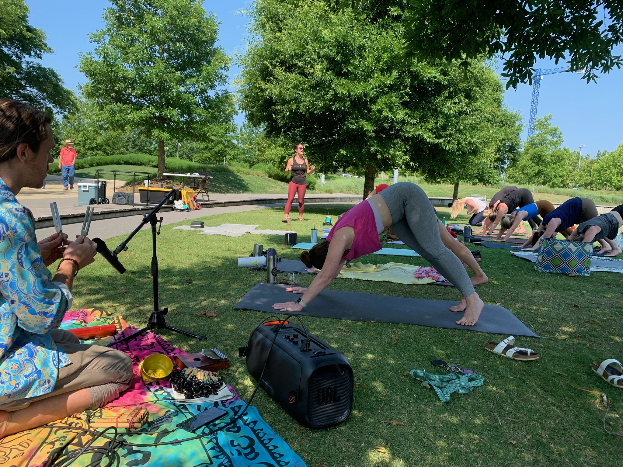 Group of people doing yoga on mat in a sunny park, instructor standing and guiding in the center.
