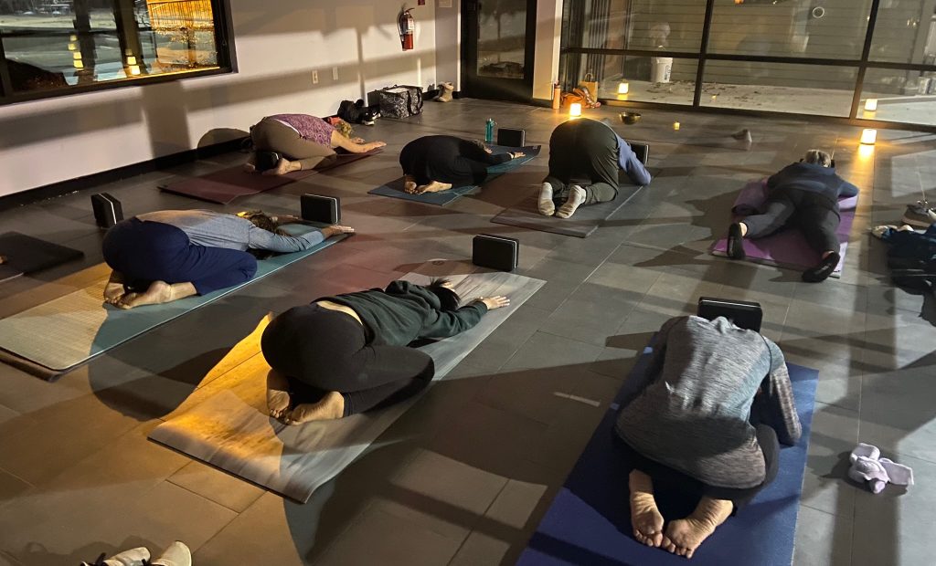 Yin Yoga in Raleigh | Unita Yoga People in a dim indoor studio practicing yoga in child's pose on mats with props and water bottles nearby, evening light through windows.